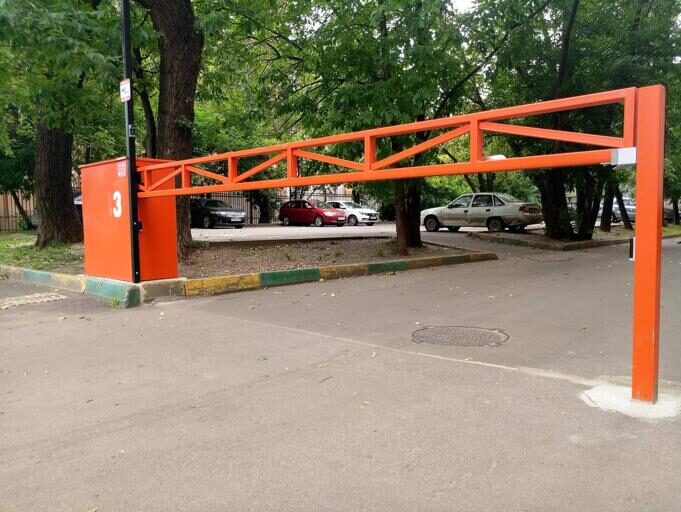 Na zdjęciu widać jaskrawo pomarańczową barierę parkingową oznaczoną numerem 3, ustawioną w pozycji podniesionej, aby umożliwić wjazd na parking otoczony drzewami, pełen zaparkowanych pojazdów. Scena rozgrywa się na tle brukowanego otoczenia, akcentowanego krawężnikami pomalowanymi na zielono i żółto.